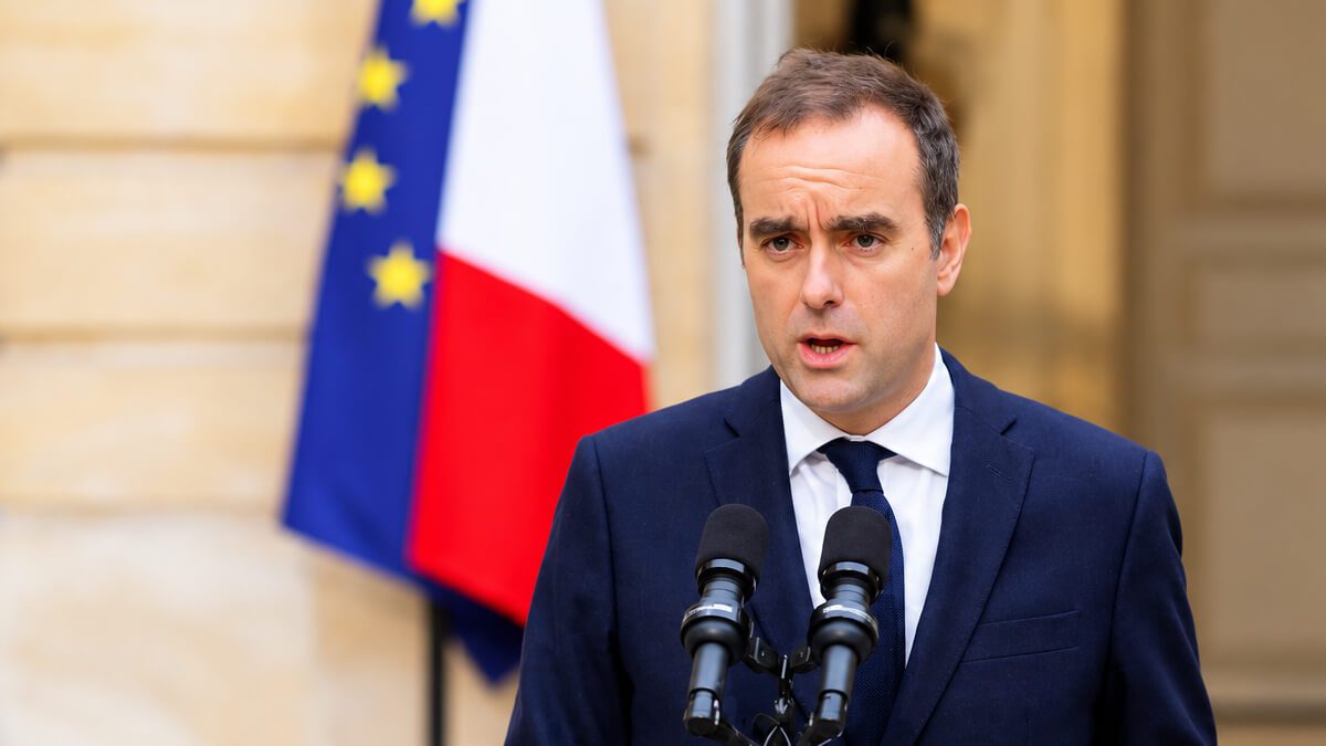 Homme en costume s'exprimant devant deux micros, drapeaux français et européen en arrière-plan